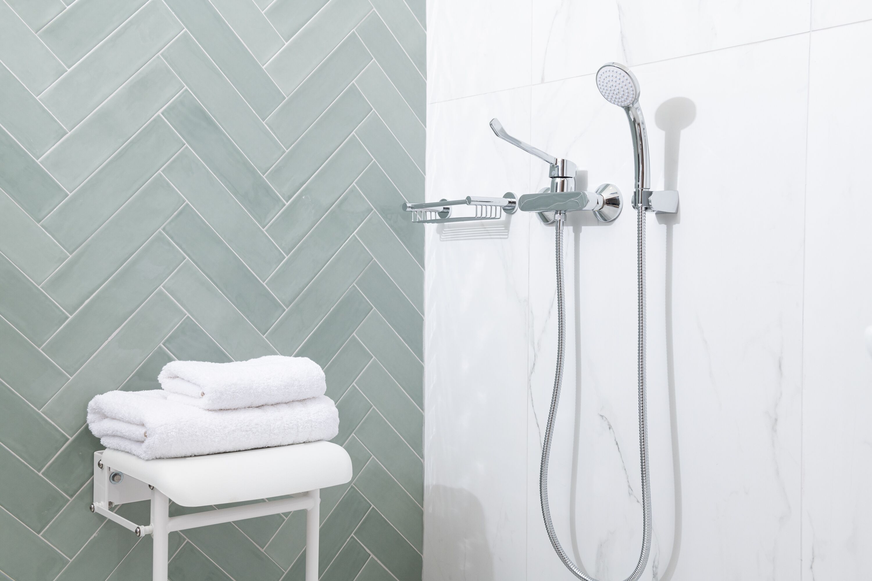 A bathroom with a herringbone tile pattern, a white bench with folded towels, and a shower with a handheld showerhead and a wall-mounted faucet.