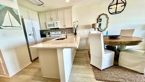 A modern kitchen with white cabinetry, a central island, and a dining area with a round wooden table and white chairs. - Luxury Gulf-Front 2BR 2BA Pet-Friendly Maravilla Resort Condo - Stunning Views (Miramar Beach)