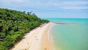 A beach with a long stretch of sand, a few people, and a line of palm trees on a hillside. - Arraial do Sol Beach Hotel (Porto Seguro)