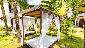 A garden with a bed under a canopy, surrounded by palm trees and a white building. - Arraial do Sol Beach Hotel (Porto Seguro)