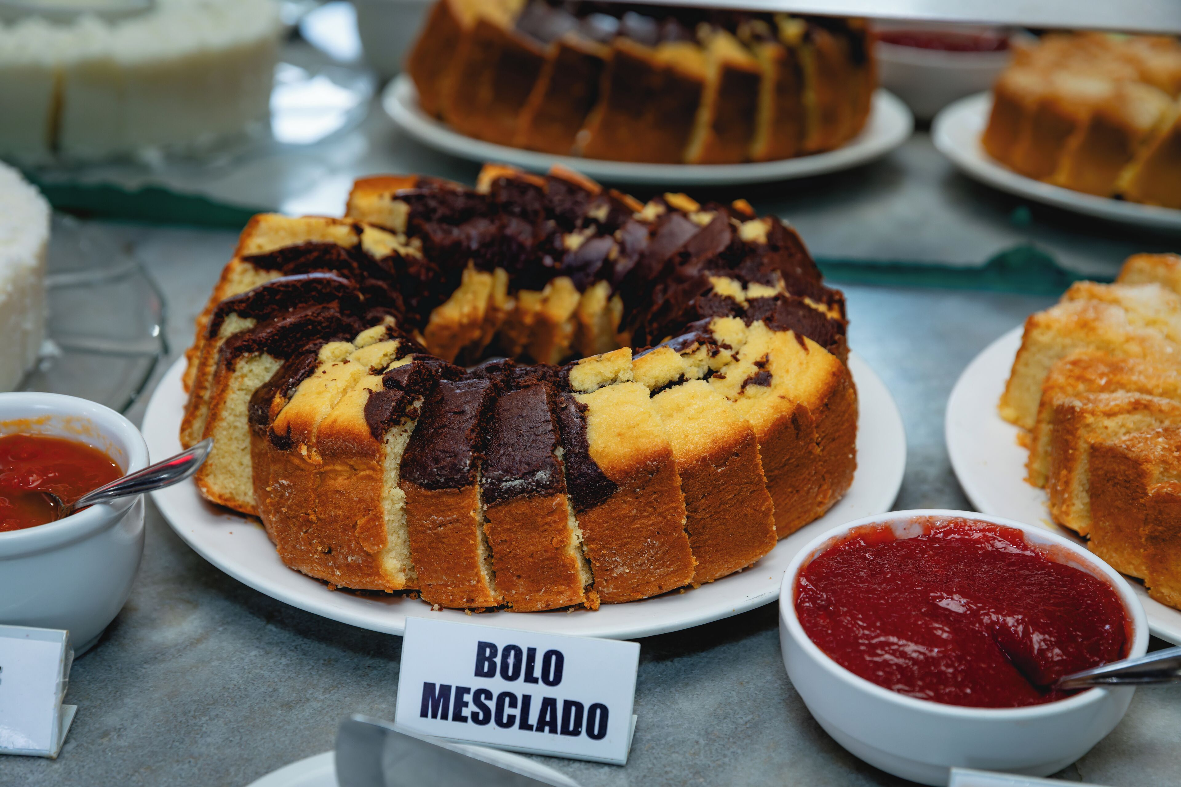 A dessert table with bolo mesclado, a chocolate cake with a yellow base, and a red sauce.
