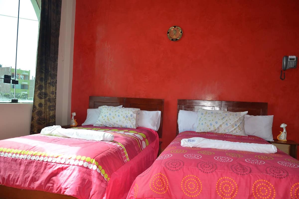 Een hotelkamer met twee bedden, elk voorzien van roze beddengoed en gestreepte kussens. Aan de muur hangt een telefoon en er is een raam met gordijnen.