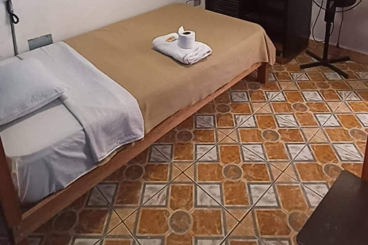 Een kleine, eenvoudige kamer met een bed, een aan de muur bevestigde televisie, een ventilator en een betegelde vloer.