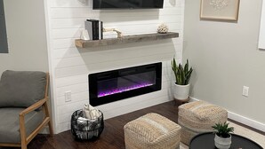 Un salon moderne avec un téléviseur à écran plat fixé au-dessus d’une cheminée, un canapé gris, deux poufs en fibres naturelles, une table basse noire et une plante en pot.