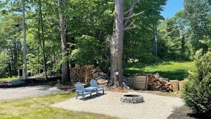 Un espace extérieur rustique aménagé avec des chaises Adirondack, un foyer en pierre et une pile de bois de chauffage.