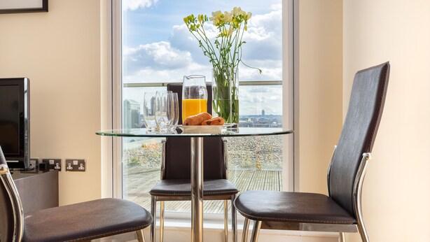 Una camera moderna con un tavolo in vetro, due sedie, un vaso con fiori, un decanter di succo d'arancia e vista sulla città attraverso ampie finestre.