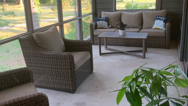 Eine überdachte Terrasse mit Korbmöbeln, einem Deckenventilator und großen Fenstern mit Blick auf eine grüne Landschaft.