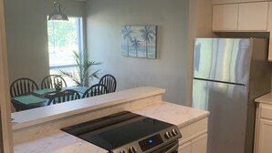 Eine Küche mit Einbauherd, Backofen und Kühlschrank. Ein Esstisch mit Stühlen und ein Fenster mit Jalousien sind ebenfalls vorhanden.