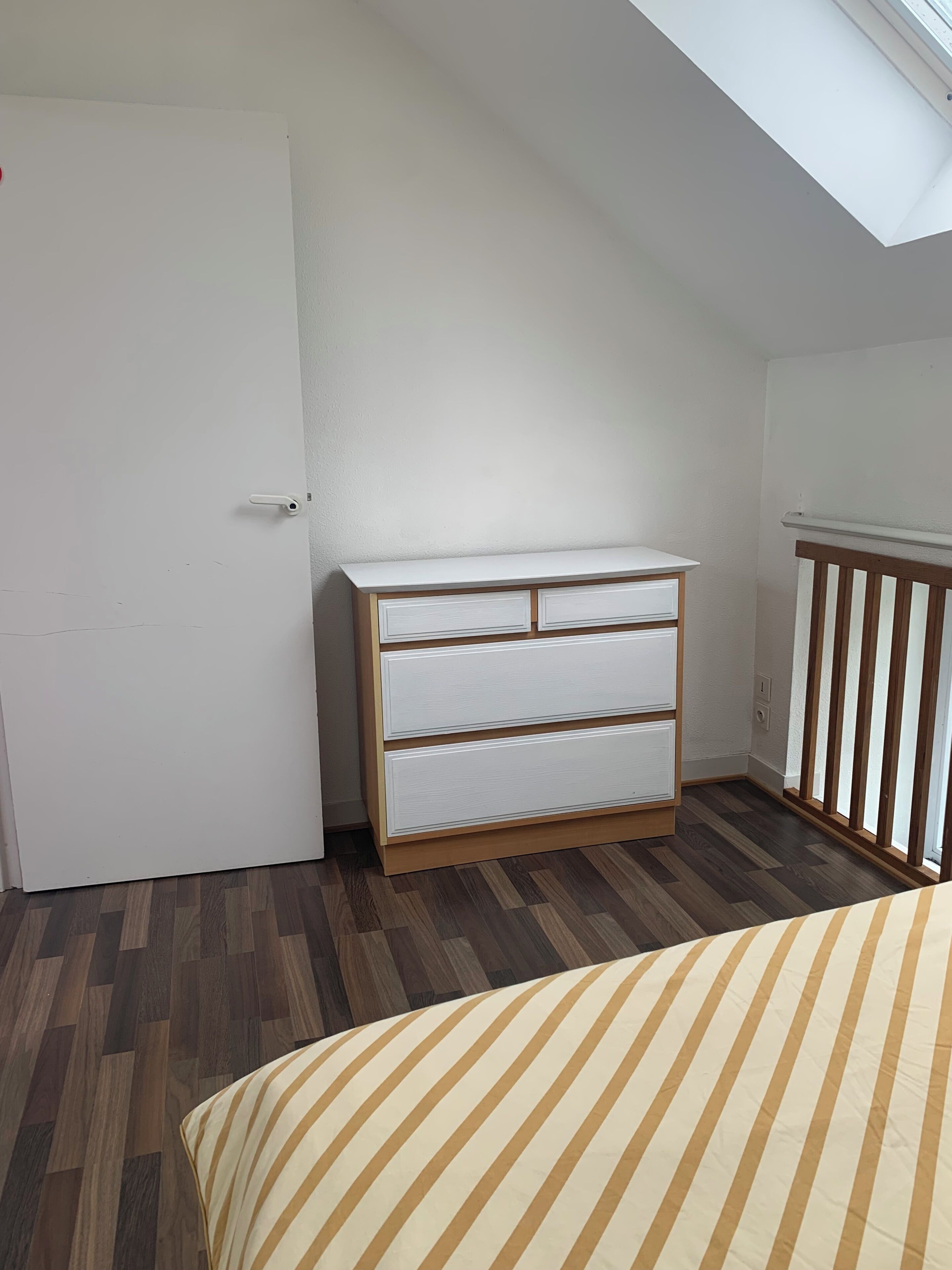 Un dormitorio con un aparador de madera, una cama con ropa de cama a rayas, un tragaluz y una puerta blanca.