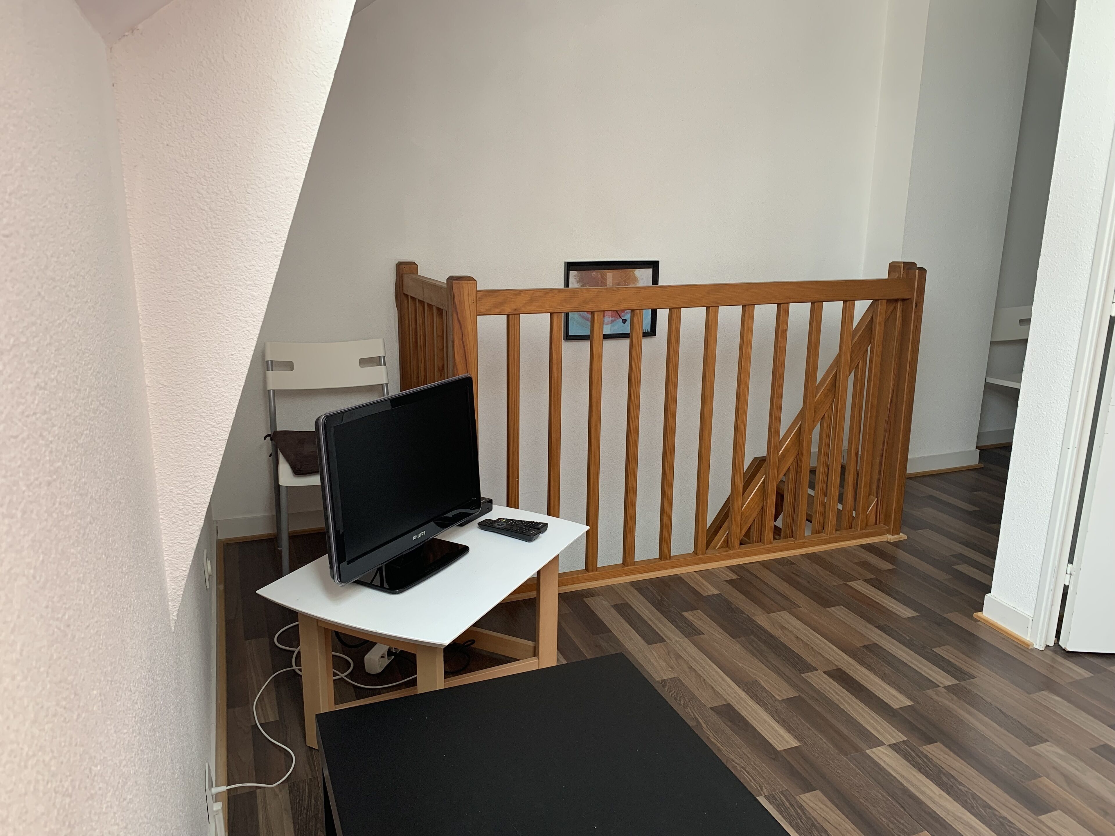 Ein Zimmer mit einem weißen Schreibtisch, einem schwarzen Monitor und einem Smartphone. Im Hintergrund ist ein Holzhandschiene und eine Treppe zu sehen.