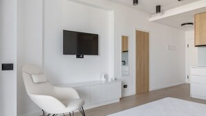 A modern hotel room with a bed, a white armchair, a flat-screen TV mounted on the wall, a wooden door, and a white cabinet. - Deluxe King Studio (Constanța)