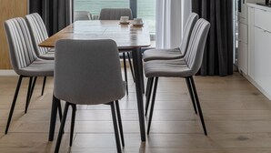 Un espace repas moderne avec une table en bois et des chaises à revêtement gris, de grandes fenêtres avec des rideaux et une vue sur la mer.