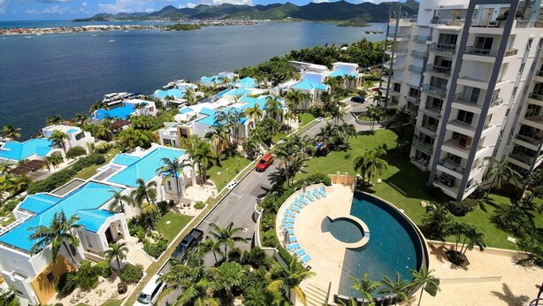 Ein Resort mit mehreren Gebäuden mit blauen Dächern, einem zentralen Schwimmbecken und Blick auf das Meer und die Berge.