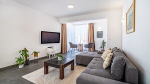 Ein modernes Wohnzimmer mit einem Flachbildfernseher, einem grauen Sofa und einem Couchtisch. Üchtitöpfe und ein großes Fenster mit Vorhängen sind ebenfalls vorhanden.