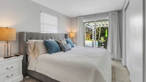 A bedroom with a large bed, a nightstand with a lamp, and a view of an outdoor pool area. - Little Beach House w/ POOLandSPA - Vanderbilt Beach (Naples)