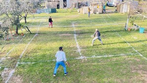 Un campo da tennis con rete al centro, circondato da un’area verdeggiante con alberi e alcuni edifici sullo sfondo.