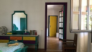Camera da letto con un letto, una cassettiera, uno specchio, una ventola e una porta che dà su un'altra stanza.