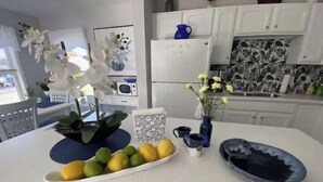 Uma cozinha com armários brancos, uma geladeira branca, um jarro azul e um vaso com flores amarelas.
