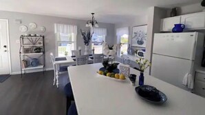 Cozinha moderna com armários brancos, uma ilha central com frutas e um vaso, e uma área de jantar com cadeiras e mesa brancas.