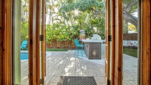 A patio with a grill, chairs, and a pool in the background. - Sunny Palms Villa - Heated Pool + Hot Tub, 2 miles to the Beach! (Fort Lauderdale)