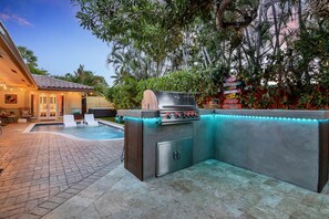 An outdoor kitchen with a grill and built-in lighting, surrounded by a pool and lush greenery. - Sunny Palms Villa - Heated Pool + Hot Tub, 2 miles to the Beach! (Fort Lauderdale)