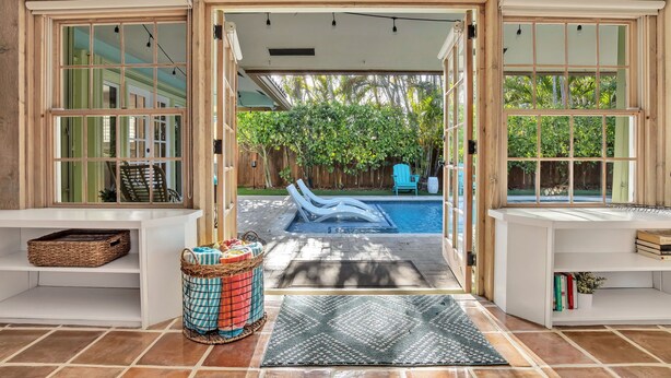 A view through a glass door into a backyard with a swimming pool, lounge chairs, and a wooden fence. - Sunny Palms Villa - Heated Pool + Hot Tub, 2 miles to the Beach! (Fort Lauderdale)