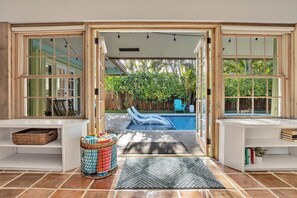 A view through a glass door into a backyard with a swimming pool, lounge chairs, and a wooden fence. - Sunny Palms Villa - Heated Pool + Hot Tub, 2 miles to the Beach! (Fort Lauderdale)