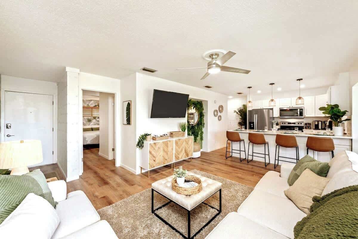 A modern living room with a sofa, a coffee table, and a TV. There is a kitchen area with a fridge, microwave, and bar stools.