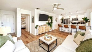 A modern living room with a sofa, a coffee table, and a TV. There is a kitchen area with a fridge, microwave, and bar stools.