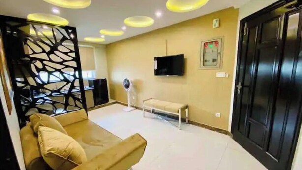Een moderne kamer met een beige bank, een ventilatorkachel, een aan de muur bevestigde televisie en een zwarte deur.