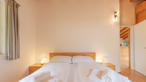 A large bed with white linens and towels, wooden headboard, bedside lamps, and a view into the room.