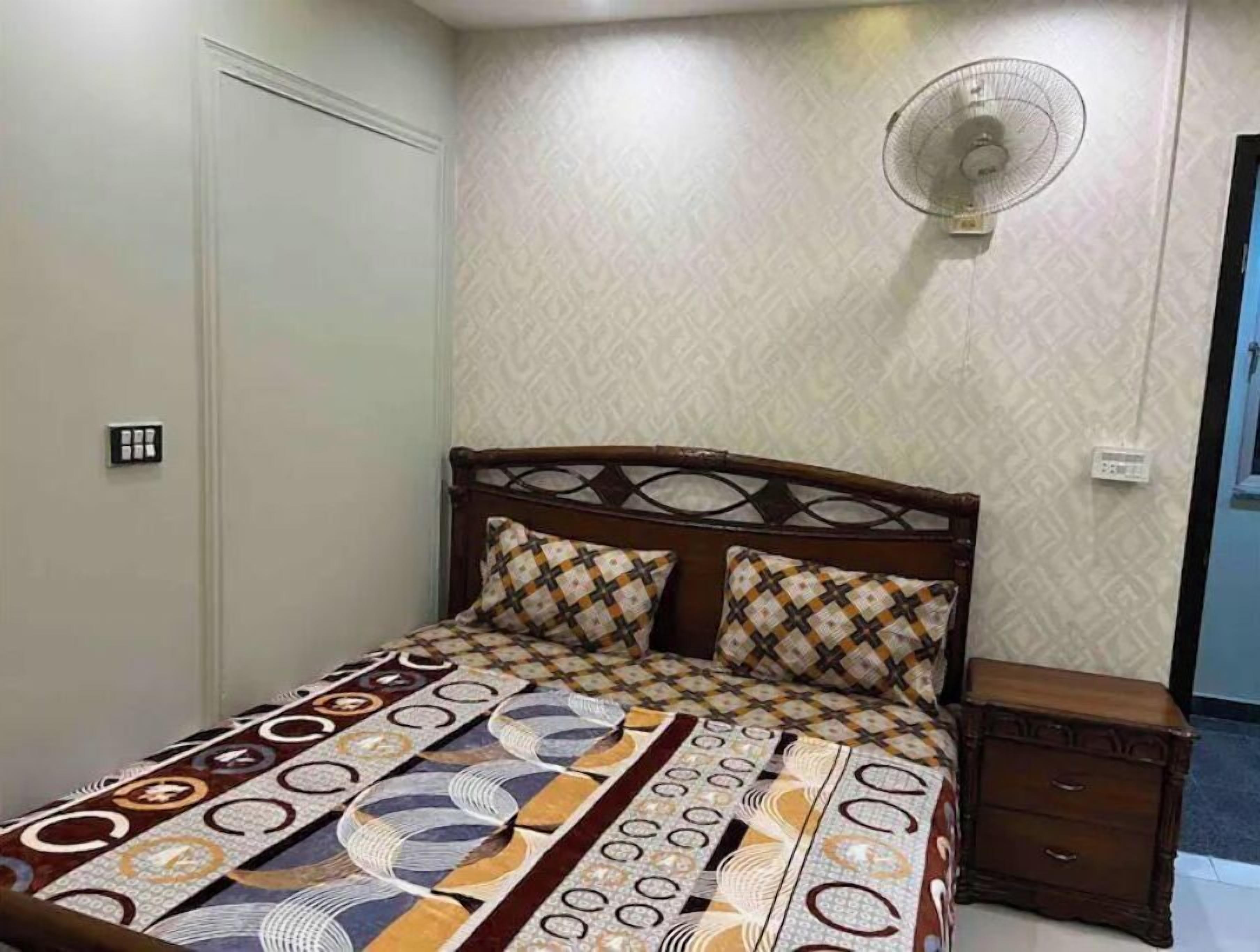 A bedroom with a bed, a nightstand, a wall-mounted fan, and patterned wallpaper.
