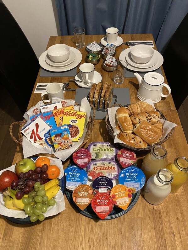 Ein Frühstückstisch ist gedeckt mit Tellern, Tassen, Brot, Gebäck und verschiedenen Frühstücksartikeln, darunter Müsli, Joghurt und Obst.