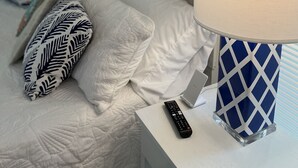 A neatly made bed with white bedding and patterned pillows, a bedside table with a remote control, and a lamp with a geometric design.