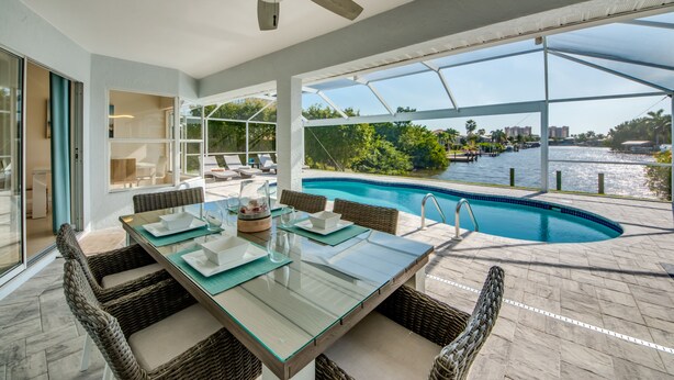 Un espace repas extérieur moderne, doté d’une table en verre et de chaises en osier, offrant une vue sur une piscine et sur le bord de l’eau.