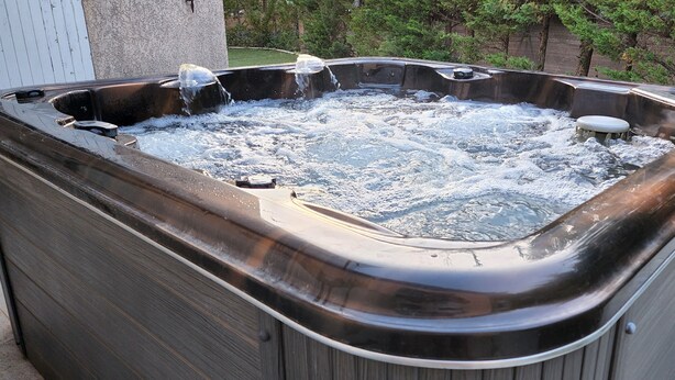 Un jacuzzi con varios chorros, rodeado por una terraza de madera y vegetación.
