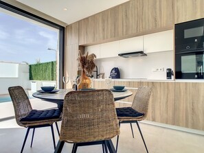 Une cuisine moderne avec des meubles en bois, un coin repas avec des chaises en osier et une table ronde, et une vue sur la verdure à l’extérieur.