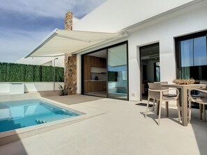 Une terrasse extérieure moderne avec une piscine, un coin repas avec des chaises et une porte vitrée donnant sur une cuisine.