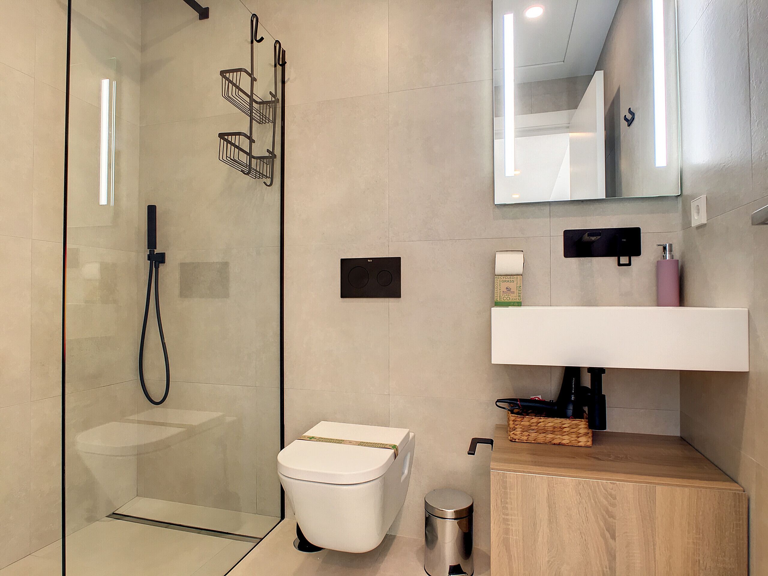 A modern bathroom with a glass shower enclosure, a toilet, a sink with a mirror above, and a trash can.