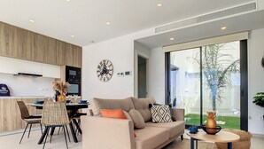 A modern living room with a sofa, a dining table, and chairs. There is a kitchen area with cabinets and a microwave. A large clock is on the wall, and there is a window with a view of a garden. - Villa with private roof terrace, patio with pool (Santiago de la Ribera)