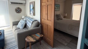 A living room with a sofa, a small wooden table, and a sliding door leading to a bedroom with a bed. -  Starfish Cottage - Summer fun.  Sleeps 4 -boating fishing and Lobster 🦞 season. (Summerland Key)