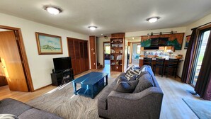 A spacious living room with a sofa, a coffee table, and a television. There is a kitchen area with a dining table and chairs visible through an open doorway. - Spacious home with large yard and hot tub- Nearby beach access and dog Friendly! (Brookings)