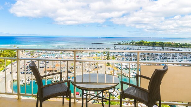 Un balcon agrémenté de fauteuils en osier et d’une table, offrant une vue sur une marina avec des bateaux et un ciel bleu azur.