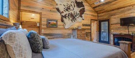 Un dormitorio de cabaña de troncos con una cama grande, paredes de madera, un trofeo de caza colgado, chimenea y televisor.