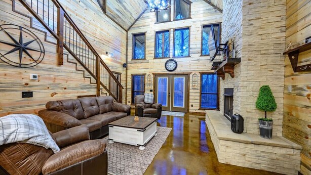 A spacious living area with a leather sofa, a coffee table, and a fireplace. A staircase leads to an upper level with large windows. - Driftaway Lodge | Luxury Broken Bow Cabin with Game Room, Hot Tub & Fire Pit (Broken Bow)