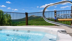 Un jacuzzi con barandilla metálica, una terraza de madera y vista a colinas verdes y una montaña al fondo.