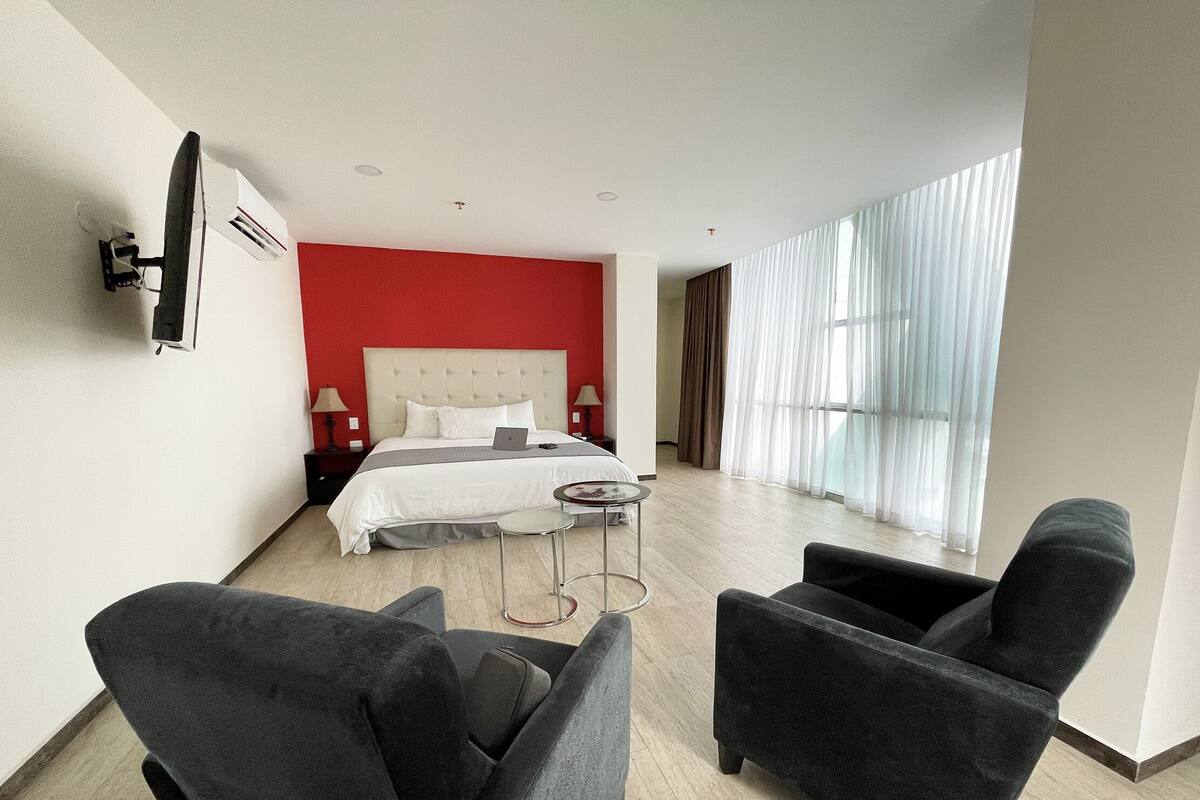 A modern hotel room with a large bed, a desk, two armchairs, and a large window with curtains.
