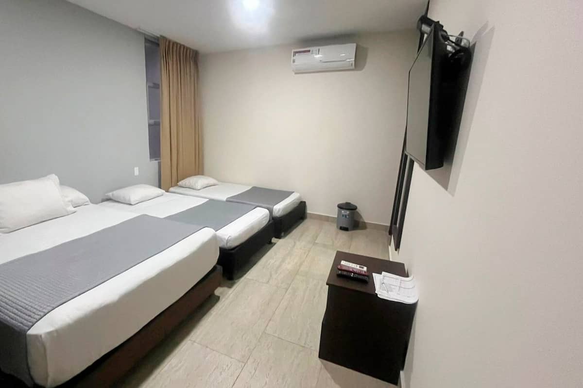 A hotel room with two beds, a small table, a trash bin, an air conditioning unit, and a television mounted on the wall.