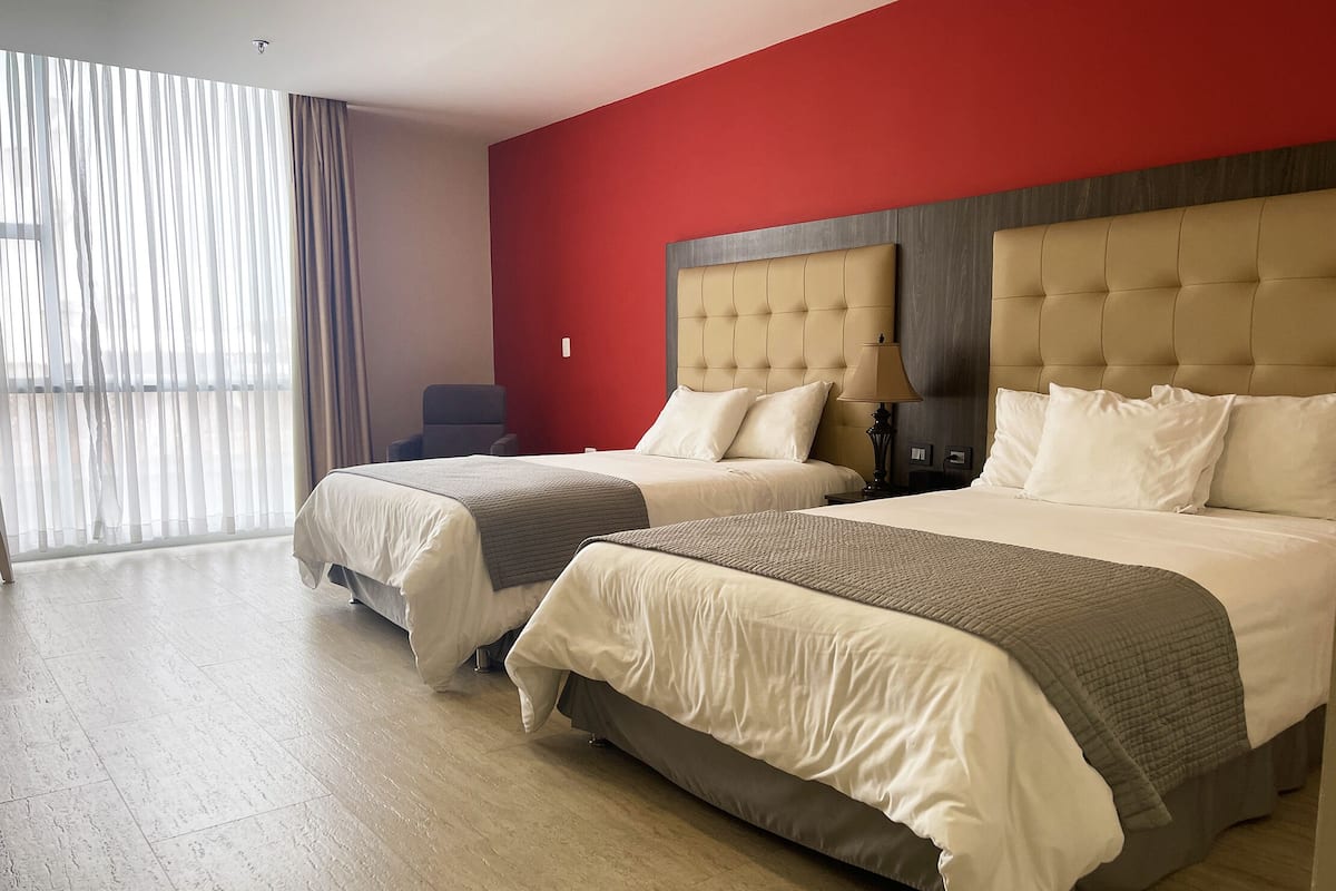 A hotel room with two beds, a red accent wall, and a large window with sheer curtains.