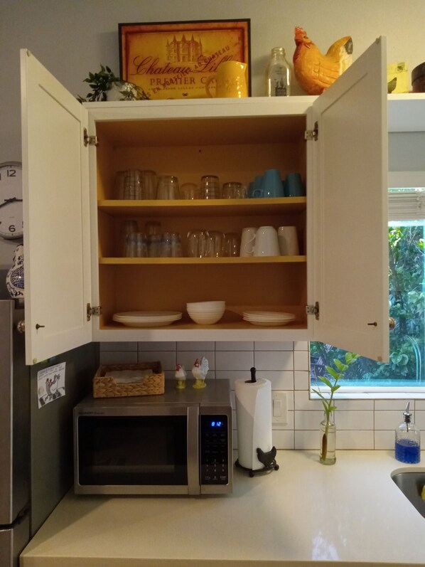 A kitchen with a microwave, open cabinet shelves with dishes, a framed picture, and a window with a view of greenery.